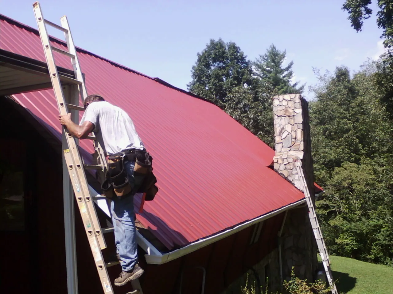 Roofer on a ladder beside a red metal roof and stone chimney — Soffit & Fascia Repair by Roofing Brothers in Armada